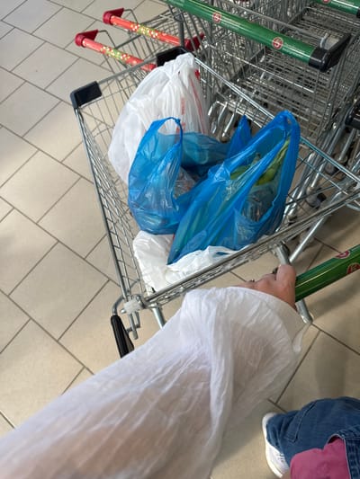 The media file shows a close-up, overhead view of shopping carts at a grocery store in Ekoposelok, Russia. The primary subjects are several connected shopping carts, some filled with plastic bags, and a person's hand and arm, clad in a white, possibly disposable, sleeve or garment. The shopping carts appear to be in a store aisle, indicated by the tiled floor and the presence of other carts in the background.

The shopping carts are made of metal and have green handles with visible text that appears to be in Russian. The text on the green handles reads "Пятёрочка выручает!" which translates to "Pyaterochka helps out!". The carts contain several blue and white plastic bags, suggesting recent shopping activity. One cart in the foreground has red and white decorative elements on its lower supports.

The hand visible is gripping the handle of one of the shopping carts. The person is wearing blue jeans and white sneakers. The white garment on the arm is somewhat translucent and crinkled, possibly a protective cover or a light jacket.

The scene appears to be indoors, under artificial lighting, with no indication of the time of day or weather. The overall impression is one of everyday commerce and personal shopping. The image focuses on the detail of the shopping cart contents and the person's interaction with the cart.