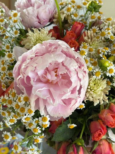 This image is a close-up, overhead shot of a vibrant bouquet of flowers. The prominent subject is a large, fluffy pink peony with delicate, layered petals. Surrounding the peony are numerous small white daisies with yellow centers, some small coral-colored roses, and a few pale yellow chrysanthemum-like flowers. The flowers are arranged in a lush, full bouquet with visible green leaves and stems. The background is blurred, suggesting the bouquet is the sole focus. No specific location in Glendale, United States of America, can be determined from the image. There are no people or actions depicted.