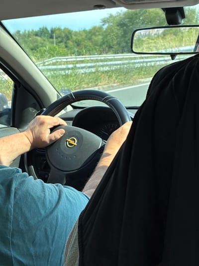 The image depicts the interior of a car, with a person driving and another passenger in the foreground. The driver's hands are visible on the steering wheel, which features an Opel logo. The car is on a road, with trees and vegetation visible through the windshield and side windows. The scene appears to be in a rural or suburban area. The passenger is wearing a black garment that covers most of their body. The image does not provide enough information to determine the specific location in Aheloy, Bulgaria.