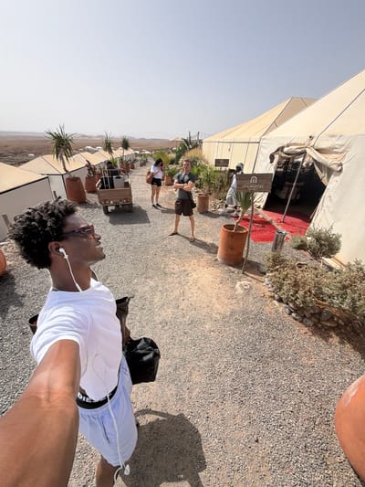 The image depicts a scene at a desert camp in Agafay, Morocco. In the foreground, a man with dark curly hair, wearing sunglasses, a white t-shirt, and light blue shorts, is taking a selfie. He has earphones in his ears and is carrying a black bag over his shoulder. Behind him, a dirt path is visible, lined with gravel.

In the mid-ground, several large, beige canvas tents are set up, suggesting accommodation. One tent has its entrance open, revealing a red carpet inside and some tables and chairs. Signs with the logo "MIA AGAFAY RESORT" are placed near the tents. A small utility vehicle with luggage is parked near one of the tents, and several other people are seen walking around the camp.

The environment is arid and desert-like, with sparse vegetation, including potted plants and bushes. In the background, a vast desert landscape stretches towards the horizon under a clear, bright sky. The overall impression is that of a desert glamping or resort experience.