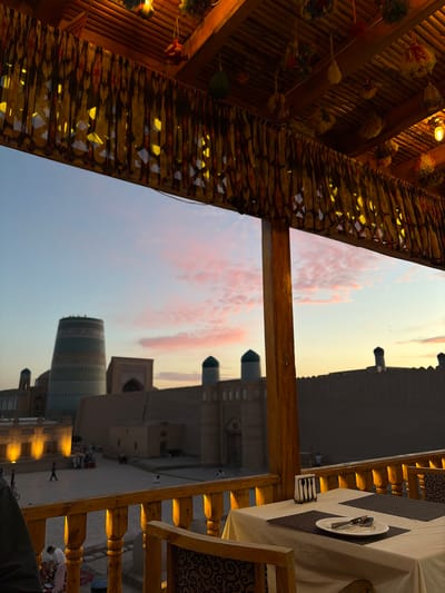 The image depicts an exterior view of a restaurant seating area overlooking a historic courtyard in Khiva, Uzbekistan, at dusk. In the foreground, a wooden railing encloses a dining table set with a white tablecloth, placemats, and cutlery. A chair with a patterned fabric is visible to the right. Beyond the railing, a courtyard with people, some sitting on rugs and others walking, is illuminated by warm lighting. Dominating the background are several ancient structures, including a tall, cylindrical minaret adorned with intricate tilework, and a fortified wall. The sky is a gradient of pale blue to soft pink, with wispy clouds indicating the setting sun. The restaurant's wooden roof structure is visible at the top of the frame, decorated with fabric and tassels.