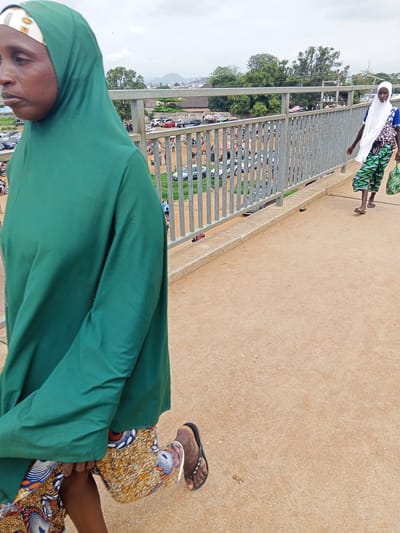 In the city of Sauka, Nigeria, on an overcast day, a woman in a green hijab and patterned skirt walks across a pedestrian bridge. She is in the foreground and is captured from the waist down. Her left leg is bent at the knee, and she is wearing flip-flops. In the background, another woman wearing a white hijab and a patterned skirt walks in the same direction. She is carrying a green bag. Below the bridge, a busy street with cars and market stalls is visible. The bridge itself has a metal railing and a concrete walkway. The overall scene depicts a typical urban environment in Nigeria.