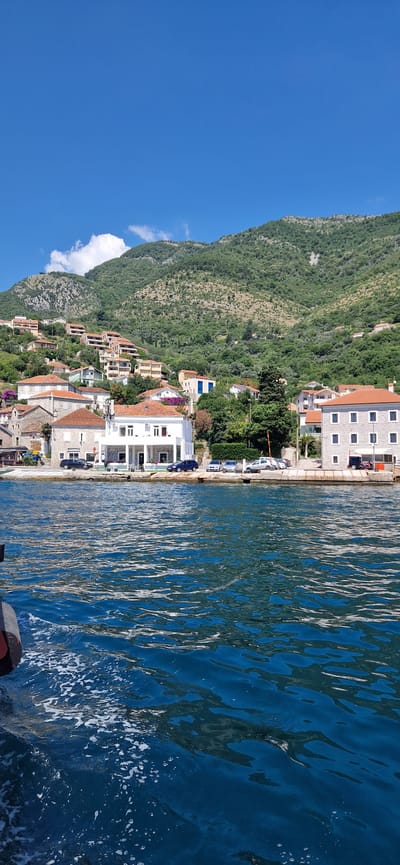 The image depicts a waterfront scene in Đurići, Montenegro. The foreground is dominated by the choppy, deep blue water of a bay or sea, with white foam trails indicating movement, possibly from a boat. In the midground, a paved walkway lines the waterfront, with several vehicles parked along it, including a dark SUV, a white sedan, and other cars partially visible. A row of buildings with terracotta tiled roofs and light-colored facades stands behind the walkway. One prominent white building features a covered outdoor seating area with tables and chairs. Further inland, more houses with similar architectural styles are nestled on a steep, verdant hillside that forms the backdrop. The hillside is covered in dense green foliage, interspersed with rocky outcrops and some scattered trees. The sky above is a clear, bright blue with a few wispy white clouds. The overall impression is one of a tranquil, sunny day in a coastal town.