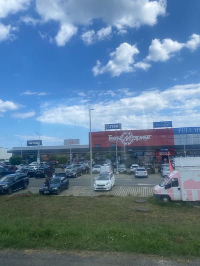 An exterior, eye-level, medium-wide shot captures a commercial area with multiple stores and a parking lot filled with vehicles under a partly cloudy blue sky. The storefronts display various brand names including "sinsay," "kik," "JYSK," "Техномаркет" (Technomarket), and "FULL HOUSE." The building facades are a mix of red and grey, with large windows. In the parking lot, numerous cars are parked, and a few people are visible walking. To the right, a pink and white van, possibly an ice cream truck, is parked. The foreground shows a grassy area with a gravel path. The scene appears to be in Ravda, Bulgaria, likely a shopping complex.