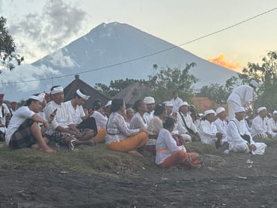 The image depicts a gathering of people sitting on a grassy slope, with a large mountain prominently visible in the background. The sky above is partly cloudy, and the sun appears to be setting, casting a warm glow. The people are mostly dressed in white, with many wearing head coverings. They are seated in rows, facing generally in the same direction, suggesting a communal event or ceremony. Some individuals are holding phones, and a few are looking at them. The foreground consists of dark, rough soil. The presence of traditional Balinese attire and the mountainous landscape strongly suggests the location is Abang, Bali, Indonesia, a region known for its volcanic scenery and strong cultural traditions. The overall atmosphere appears to be peaceful and contemplative.