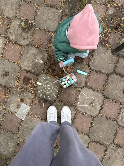 A high-angle shot shows a young child and an adult in an outdoor paved area in Tomilino, Russia. The child, wearing a pink hooded cap, a green jacket with white striped cuffs, and blue gloves, is on the left, holding a blue chalk stick and interacting with items in a small container. Beside the child is an open container of colored chalk. On the paved surface are chalk drawings: a sun with a smiley face and a heart. To the right, an adult stands with feet positioned slightly apart, wearing gray pants and white sneakers. Debris such as small twigs and fallen leaves are scattered across the pavement. A small clear plastic container and a pine cone are on the pavement near the adult's feet. The pavement is made of interlocking, flower-shaped paving stones.