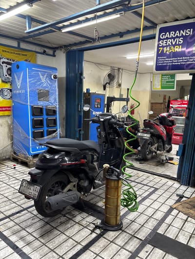 This media file captures a scene inside a vehicle service center, likely a motorcycle repair shop, in Binong, Indonesia. The focus is on two motorcycles, one black and one red, positioned for service. The black motorcycle in the foreground is partially disassembled, with its engine area exposed. A person, presumably a mechanic, is visible working on the rear tire of the red motorcycle.

Various service equipment is present, including a blue machine covered in plastic wrap with the "VINFAST" logo, a tire balancing machine, and an oil drain or collection system with a green hose. The setting appears to be indoors, with fluorescent lighting illuminating the tiled floor.

Prominent text includes a sign for "GARANSI SERVIS 7 HARI" (7-Day Service Warranty) with terms and conditions, and another sign mentioning "SERVIS RASAMIN TERBUKTI MAMPU MENURUNKAN EMISI SECARA SIGNIFIKAN!" (Rasamin Service Proven to Significantly Reduce Emissions!). There are also brand logos such as "HONDA" and "Coca-Cola". The presence of the "VINFAST" logo suggests this might be a dealership or service center for that brand as well. The overall atmosphere suggests a busy, functional workshop environment.
