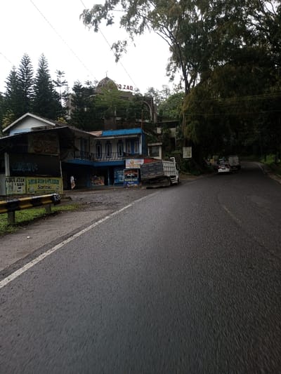 The image presents a roadside scene in the city of YonPhoogla, Bhutan, under an overcast sky. In the foreground, a dark asphalt road, appearing damp, stretches from the bottom center, curving slightly to the right as it recedes into the distance. A continuous white line marks the left edge of the road, adjacent to a dirt and gravel shoulder. A yellow and black striped metal safety barrier runs along the immediate left.
Behind the barrier, a cluster of buildings is visible. The closest is a single-story structure with a dark roof and a blue-painted lower facade, featuring several commercial signs in Indonesian text, including "UBUR ATAM U KOPI", "BUBUR AYAM & MIE REBUS Sarasa", and "WARUNG MAM". A single individual, dressed in light-colored clothing, stands near the entrance of this building. A white dump truck with a dark, open bed is parked partially on the dirt shoulder and partially on the road directly in front of these buildings, facing away from the viewer.
Further back and uphill, behind the immediate structures, a larger, multi-story building with a domed roof and the text "GANG ZAQ" visible on its facade is partially obscured by trees, including several tall evergreen conifers. Dense foliage and tall trees line the right side of the road, extending into the background. In the mid-distance on the right side of the road, a white sedan and a dark-colored truck are parked on the shoulder, both facing away. Overhead, faint power lines are discernible against the light gray sky.