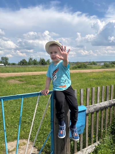 A young Caucasian child wearing a blue t-shirt with a graphic print and black pants sits on a rustic wooden fence. The child has on a straw hat and blue sandals with rocket designs. The child's right hand is raised, palm facing the camera. The background features a grassy field with a dirt road, trees, and a few houses in the distance under a cloudy blue sky. The scene appears to be in a rural area, possibly on the outskirts of the city of Kurgan, Russia. A blue mailbox is attached to the fence near the child.