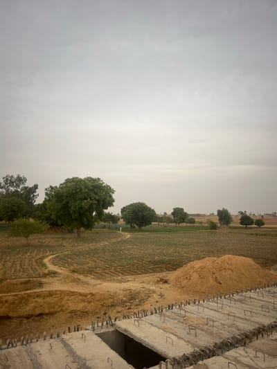 A rural landscape fills the frame, a cloudy sky dominating the view. Green fields stretch out, dotted with clusters of trees. A dirt path weaves through the fields, leading into the distance. In the foreground, a raw concrete structure, possibly a building under construction, sits partially in the frame. Protruding metal bars reinforce the concrete. Adjacent to the structure, a small pile of dirt sits near the edge of the field.