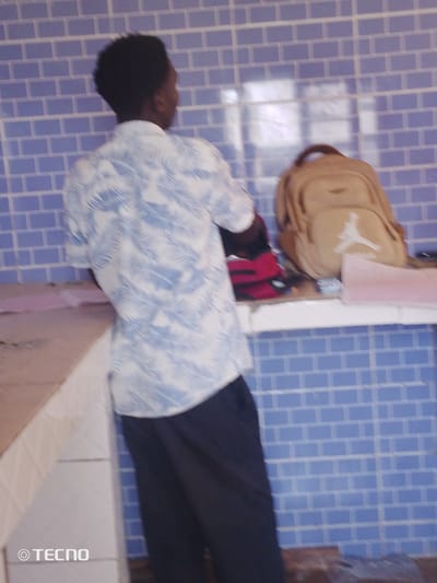 A person, seen from behind, stands facing a blue tiled wall in a room. They are wearing a white short-sleeved shirt with a blue leaf pattern and dark pants. To their right, on a shelf, is a tan backpack with a white Jordan logo, and a red object, possibly another bag. The room has a counter to the left. The overall impression is of a domestic or casual setting within Gumel, Nigeria.