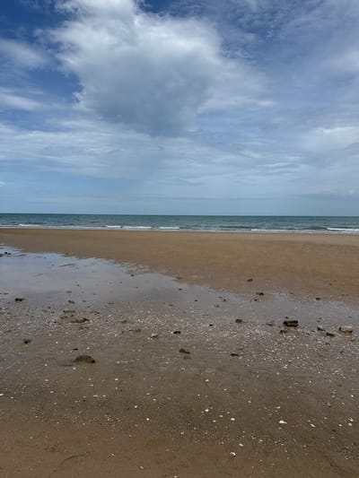 The image shows a wide, sandy beach with calm, blue-green ocean waves gently lapping the shore. The sky is filled with a mix of white and gray clouds, suggesting an overcast or partly cloudy day. Scattered across the wet sand in the foreground are small shells and pebbles. The scene depicts a natural coastal environment. This location is identified as Ban Bang Ket, Thailand.
