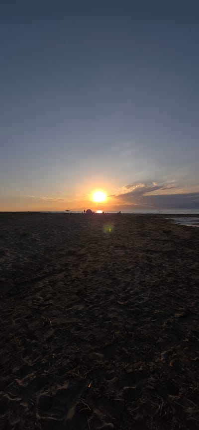 The image captures a serene sunset over a beach in Boz Beshik, Kyrgyzstan. The sky is a gradient of deep blue at the top transitioning to lighter hues near the horizon, with soft clouds tinged orange by the setting sun. The sun itself is a bright, radiant orb positioned just above the distant horizon, casting a warm glow across the scene.
In the middle ground, a stretch of dark sand forms the beach. Scattered across it are several small groups of people, appearing as silhouetted figures against the bright sky. Some individuals seem to be standing, while others might be seated or engaged in activities near what appears to be a beach umbrella and perhaps a tent. The sand itself is textured, showing footprints and small debris, suggesting recent activity. To the right, a calm body of water is visible, reflecting the colors of the sunset. The foreground is dominated by the dark, textured sand, drawing the viewer's eye towards the illuminated horizon.