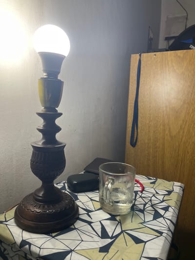 A lit lamp sits on a patterned surface in Bernal Este, Argentina. A glass of water and a black rectangular object are nearby. A wooden surface is above and to the left of the lamp.