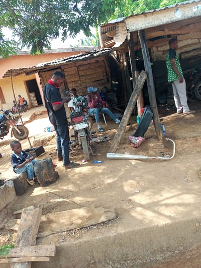 This image depicts an outdoor scene in Aramoko-Ekiti, Nigeria, functioning as a motorcycle repair or social gathering area under direct sunlight. The ground is unpaved, consisting of dry, reddish-brown dirt, with a concrete curb and blocks in the foreground.

In the center, a male individual in a black t-shirt and dark pants stands, examining a small object held in his hands, positioned directly over a partially disassembled motorcycle with a visible Nigerian license plate (GA...48B). To his immediate left, a young boy in a blue striped polo shirt and blue jeans is seated on a concrete block, also focused on an object in his hands.

Behind the central figures, a rudimentary structure with walls of rough wooden planks and a corrugated metal roof provides shelter, supported by thick wooden beams. Within this structure, two men are seated: one in a blue baseball cap, red jacket, and blue jeans, with a hand raised towards his face, possibly using a phone; another less distinct male figure is partially visible behind him. To the right, within the same structure, a third man in a green patterned t-shirt and light pants stands, facing right.

Additional objects include a silver motorcycle exhaust pipe lying on the ground in the mid-right foreground, a black and pink motorcycle seat leaning against a wooden beam, and various small components scattered. Another motorcycle is partially visible in the left background near a light peach-colored building with a corrugated roof, in front of which a group of several children are present. Green foliage from overhead trees casts distinct shadows across the scene.