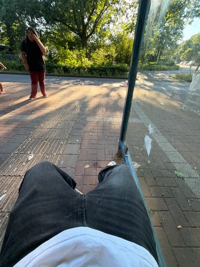 The image depicts a view from a person's seated perspective at a bus stop, likely in Dorsten, Germany, given the context. The person is wearing dark jeans and a white shirt. In the background, a woman in a black t-shirt and maroon pants stands to the left, looking away and touching her face. The bus stop structure features a transparent glass panel, reflecting the surrounding greenery and sunlight. The ground is paved with bricks and scattered with cigarette butts. The environment appears to be an outdoor, urban setting with trees and sunlight creating dappled shadows on the ground.