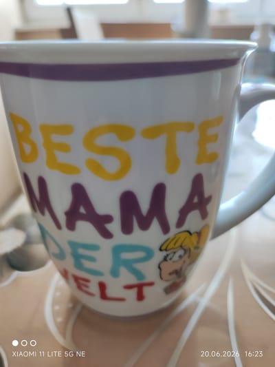 The image displays a white ceramic mug with colorful lettering and a cartoon character. The mug has a purple band near the rim and a handle on the right side. The lettering on the mug reads "BESTE MAMA DER WELT" in yellow, purple, turquoise, and red. A cartoon depiction of a blonde-haired girl with a large smile and wide eyes is on the lower right side of the mug. The mug is placed on a surface with a patterned texture, possibly a table. In the background, out of focus, are hints of indoor elements like windows and furniture. The timestamp in the bottom right corner indicates "20.06.2026 16:23" and the text "XIAOMI 11 LITE 5G NE" is in the bottom left corner, which refers to the phone used to take the photo. There is no indication of the city of Böblingen, Germany in the image itself.