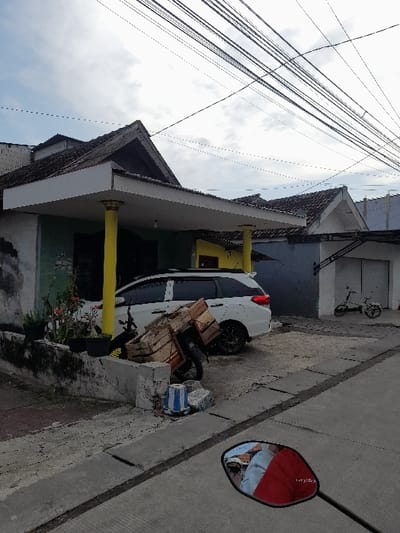 The scene is a residential street in Kecamatan Tanggulangin, Indonesia, located at latitude -7.506, longitude 112.723. A white SUV is parked in front of a house with a yellow awning and a wooden cart beside it. The house has a yellow pillar and a green wall with a small plant in front of it. Another house, with a gray wall, is seen in the background. A blue cylindrical object is on the ground, along with other debris. A black motorbike is partially visible in the foreground, with its rearview mirror reflecting a person wearing a red shirt. Numerous power lines are visible overhead. The street is paved and appears to be empty.