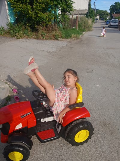 A young girl with dark hair, wearing a floral sundress and pink sandals, is sitting on a red toy tractor. She is positioned on the tractor's body, with her legs extended and her feet resting on the ground. A yellow seat is visible behind her. The tractor has large black tires with yellow rims. A pink child's shoe is on the ground near the tractor.
The scene is an outdoor setting with an asphalt road. To the left of the road, there are green trees and bushes, as well as a white building. In the background, a white van and a blue car are parked on the road, and a person is walking in the distance. The time of day appears to be daytime, likely late afternoon, given the sunlight casting shadows. The weather seems to be warm and sunny.
The visible text on the tractor reads "FASTRAC". The overall mood of the image appears to be playful and carefree, suggesting a child enjoying an outdoor activity.