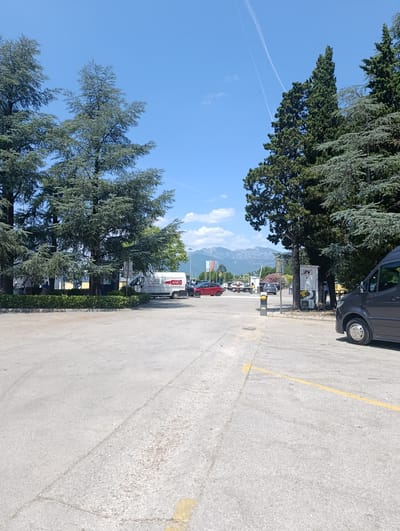 The image depicts an outdoor scene on a bright, sunny day, likely in the city of Mrčevac, Montenegro. In the foreground, a grey van is parked on an asphalt lot marked with yellow lines. To the left, several large evergreen trees with feathery foliage are prominent, with their branches reaching towards a clear blue sky. Further into the midground, there is a white van with "LANEX" branding visible on its side, parked in front of a low green hedge. A few people are seated near the hedge. Beyond the vans, more trees are visible, and in the distance, a range of mountains with a hazy appearance can be seen under a sky with faint contrails. Other vehicles, including a red car and several dark-colored cars, are parked or driving in the midground, suggesting a public area or parking lot. A modern-looking payment or service booth with a digital display is situated to the right of the white van. The overall environment suggests a commercial or public parking area, possibly near a business or attraction, with a backdrop of natural mountainous scenery.
