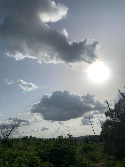 The image, taken from a slightly low angle, shows a partly cloudy sky with the sun prominently visible. Two large cloud formations dominate the left and center of the frame. The left cloud is lighter, while the central one is darker and denser. Smaller, scattered clouds appear in the sky.
In the background, on the right side of the image, dense green vegetation and trees are visible. A few buildings can be discerned amongst the foliage. Power lines stretch across the sky, intersecting with the trees.
The location is identified as Gbeleko, Nigeria. There are no people visible in the image.