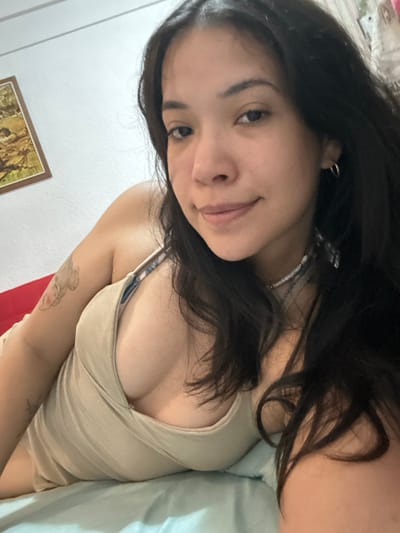 The image displays a female individual, appearing to be a young adult, with long dark hair and a light complexion. She is lying down, angled towards the camera, capturing a selfie, and exhibits a faint smile. She is wearing a light beige/tan low-cut top with thin straps. Multiple necklaces are visible around her neck, including one composed of white beads or shells and another thinner metallic chain. Small silver hoop earrings are worn. A light-colored tattoo, depicting what appears to be a butterfly, is visible on her right upper arm. The background features a plain white wall, with a framed picture or artwork of indistinct content and earthy tones hanging on the wall in the upper left. A red surface, possibly a headboard or cushion, is positioned behind her. In the foreground and beneath her, a light blue/teal fabric, consistent with a bedsheet or blanket, is present. The scene takes place indoors, within what appears to be a private dwelling in the city of Mar del Plata, Argentina.