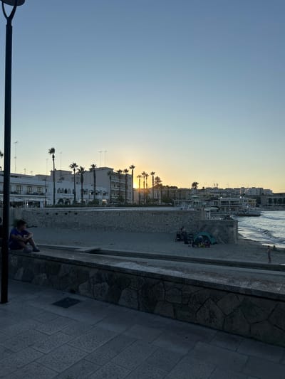 A young person sits on a stone wall overlooking a beach in Otranto, Italy. The sun is setting, casting a warm glow over the scene. In the background, palm trees line the shore, and buildings of white stone rise behind them. A few people are visible on the beach, some with tents set up. The ocean waves are gentle, and the sky is a clear, pale blue. A tall, dark lamp post stands in the foreground on the left side of the frame. The stone wall has a rough, uneven texture, and the paving stones below it are laid in a patterned arrangement.
