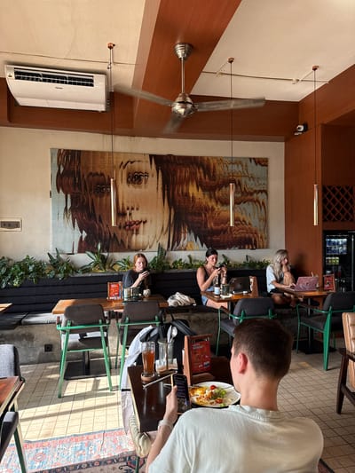 The image shows the interior of a restaurant or cafe in Uluwatu, Indonesia. Several people are seated at tables, appearing to be customers. Some are using laptops and phones, while others have food and drinks in front of them. The establishment features an eye-catching mural of a woman's face on the wall. The room is lit by pendant lights and has an air conditioner unit mounted on the wall. The dining area is decorated with greenery, and the decor includes a rug. The cafe setting suggests a relaxed, modern atmosphere.