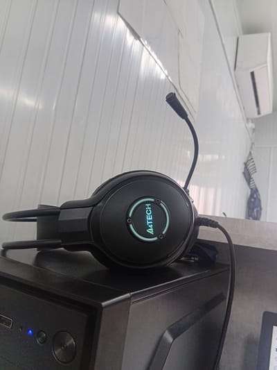 A black A4Tech headset with a flexible microphone is positioned on top of a black computer tower. The headset has illuminated green accents on its earcups, featuring the A4Tech logo. The computer tower has a blue LED light indicating it is on. The background consists of a white, ribbed wall and a light-colored ventilation unit mounted on the wall. A grey countertop is visible beneath the headset and computer tower. This scene is indoors, likely in a room or office. The location is specified as Tokmok City, Kyrgyzstan.