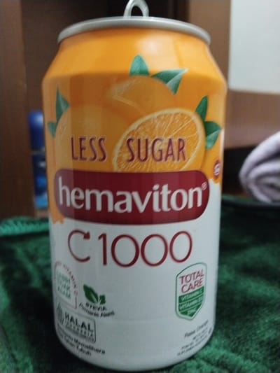 A can of Hemaviton C 1000 is depicted in a close-up, slightly angled shot. The can is primarily orange and white, featuring an illustration of a sliced orange and green leaves. Prominent text includes "LESS SUGAR," "Hemaviton," "C 1000," and "TOTAL CARE." Other smaller text elements are visible, such as "VITAMIN C," "VITAMIN B5," and "ZINC." A "HALAL" certification logo is also present. The can rests on a dark green, textured surface, possibly a cloth. The background is blurred and appears to be indoors, with hints of brown and a blue object on the left. There is no indication of the city of Lahat, Indonesia, within the image itself.