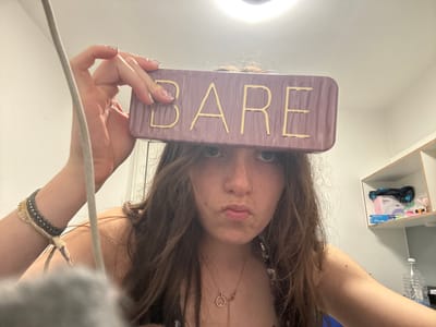A young individual, likely female, with long, wavy medium-brown hair, poses for a close-up photograph indoors. She holds a rectangular, reddish-purple object with the word "BARE" written in gold capital letters across her forehead, obscuring the upper part of her face except for her eyes. Her lips are pursed in a pout, and she has a small nose ring on her left nostril.

She is wearing a delicate chain necklace with a circular pendant containing small stones. On her left wrist, she has a multi-layered bracelet made of fabric and metal beads. Her nails are manicured with a light, possibly iridescent, polish.

The setting appears to be a simple indoor room, possibly a bedroom or dorm, indicated by plain white walls and a white shelving unit in the background on the right. On the shelves, a pink box with the brand "SUN" visible, along with other small containers and a black fabric item, can be seen. Below the shelves, a clear plastic water bottle is partially visible. A light fixture is discernible at the top center of the frame. A grey, fuzzy object and a light-colored cord run vertically along the left side, partially obscuring the scene. The lighting is artificial.