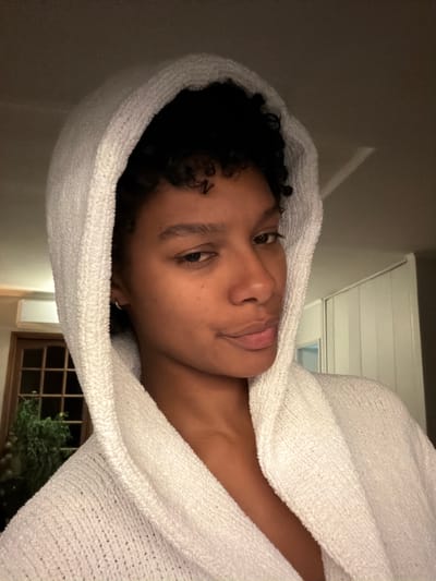 A person with short, dark, curly hair is wrapped in a white hooded towel, facing the camera. The background features a partially visible window with a view of foliage, a white ceiling, and a light-colored wall. There is no definitive information in the image to indicate the location is Champs-sur-Marne, France. The scene appears to be indoors, likely a bathroom or similar space.