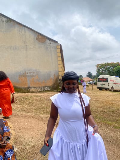 An outdoor scene in the city of Riyom, Nigeria, features a woman in a white dress in the foreground, her gaze directed downward. She wears a black headscarf and her braided hair cascades over her shoulder. In her right hand, she holds a dark smartphone, and a silver wristwatch adorns her left wrist. The background reveals an aged, neutral-toned building with signs of wear and tear. Further back, an ambulance with "AMBULANCE" written on its side is parked, suggesting a medical or emergency context. Other individuals are visible in the distance, some in white attire. The ground is covered in dry grass, and the sky is cloudy.