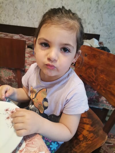 A young child sits at a table indoors, likely in a private residence, though no specific identifiers exist. The child wears a light-colored T-shirt with a graphic design. The child is wearing hoop earrings and is seated on a wooden chair. A plate is visible in the foreground. No evidence indicates the location is Pobeda, Bulgaria.