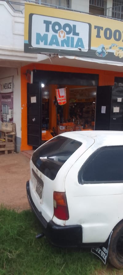 The image shows a white Toyota Corolla station wagon parked in front of a store with a sign that reads "Tool Mania." The store is located in Bungoma, Kenya, at coordinates 0.565 latitude and 34.562 longitude. The car is parked on a dirt lot, with green grass growing in the foreground. The car's rear window is tinted, and its rear bumper is black. The car has a license plate that reads "KAK 603W." The store appears to sell tools and other hardware items. The store has a large window display that shows various tools and other items. The store's door is open, and there are a few people visible inside. The environment is typical of a commercial area in Bungoma, Kenya.