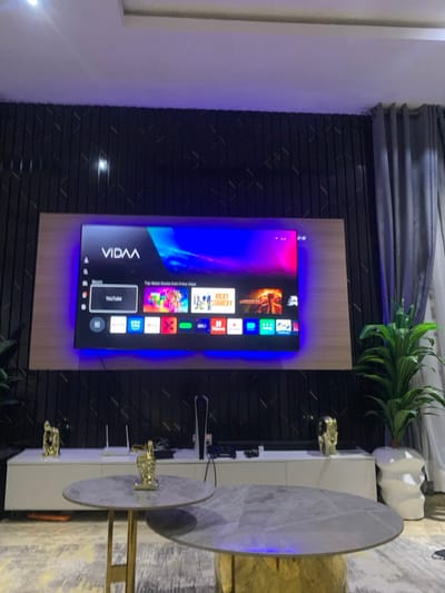 A modern living room setting features a large, wall-mounted VIDAA smart TV with a brightly lit interface displaying streaming options. The TV is bordered by a black textured wall panel and set against a light wood-toned backing. To the right, a white entertainment center houses gaming consoles, while decorative gold figurines add a touch of elegance. A dual-tiered coffee table with marble-patterned tops stands nearby, complementing the interior's stylish aesthetic. A green plant in a white pot adds a natural element.