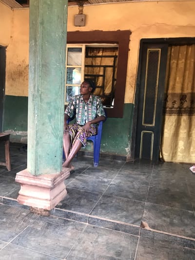 A person sits on a blue chair, leaning against a green column on a tiled floor. They wear a patterned shirt and patterned skirt. Behind them is a window with visible bars. Above, a dark door is set into a wall covered in a patterned fabric. The building's architecture shows a combination of natural materials with tiled floors, columns, and walls finished in shades of yellow and green. The setting seems to be a partially covered outdoor area.