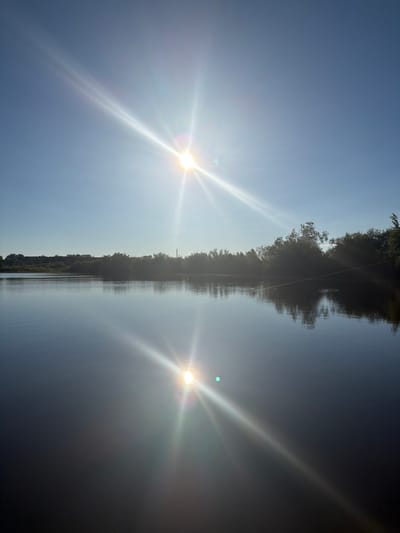 The image depicts a serene lake with a clear blue sky. The sun is prominently positioned in the upper left quadrant, casting brilliant rays of light across the scene. A reflection of the sun is visible on the calm surface of the water, creating a dazzling effect. The distant shoreline is lined with lush green trees, adding a natural beauty to the landscape. The location is identified as Tskhinevali, Russia. The scene is devoid of any people or animals, focusing solely on the natural elements and the interplay of light and water.