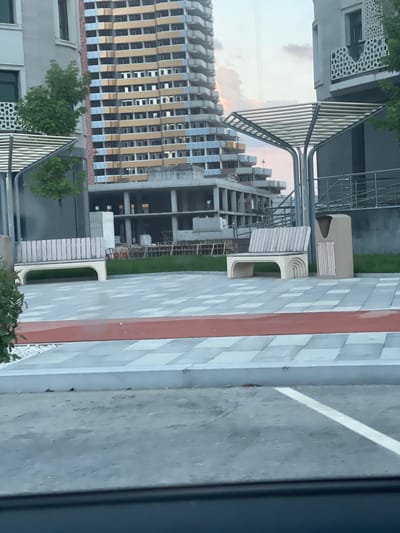 The image depicts a construction site with several buildings in different stages of completion. A tall, modern building with a facade of vertical gold and blue elements is in the foreground. Adjacent to it, another building under construction shows exposed concrete and scaffolding. The scene is set outdoors, with a cloudy sky visible in the background, suggesting it could be daytime. A paved walkway with a distinctive red and white striped pattern runs alongside the buildings. Green grass and trees are interspersed throughout the scene. Some outdoor seating or decorative elements are also visible. The image appears to be taken from a slightly elevated perspective, possibly from another building or a higher vantage point. There is no visible text in the image. The overall impression is one of urban development and ongoing construction in a modern setting.