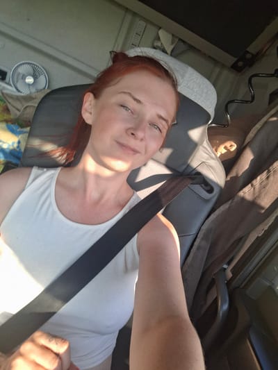 A woman with red hair and fair skin is sitting in the passenger seat of a vehicle, looking at the camera. She is wearing a white tank top and a seatbelt is across her chest. The interior of the vehicle appears to be a truck or RV, with a headrest visible behind her. A small fan is mounted on the wall to her left, and there are blankets and other personal items in the background. Sunlight streams in from the side, casting shadows across the scene. The image is a selfie, taken from a slightly elevated angle. The location is not identifiable as Kokui, Russia.