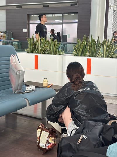This is a photograph taken inside Incheon International Airport, South Korea. The scene captures a woman from behind, sitting on a blue airport bench and reaching down towards her belongings on the floor. She is wearing a black jacket with white lettering that reads "Matin Kim" and grey shorts. Beside her, on the bench, is a shopping bag, a white camera, a phone, and a bottle of drink. On the wooden floor, near her feet, lies a crumpled bag of snacks and several black bags, one of which has its zipper prominently visible. In the background, other people can be seen walking through the airport terminal. Large potted plants with green and yellow striped leaves are positioned in planters, separating different areas of the terminal. The overall environment is clean and modern, characteristic of an airport lounge.