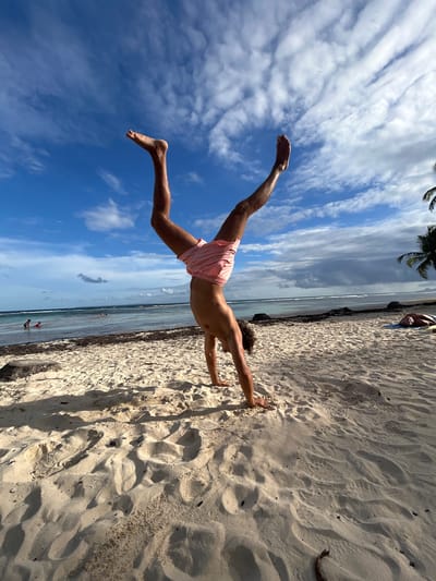 A man in pink shorts performs a plank on a sandy beach. His hands are pressed into the sand while his legs are extended upwards towards a partly cloudy blue sky. The beach is located near Sainte-Anne, France, with palm trees visible in the background, along with the ocean and other beachgoers.