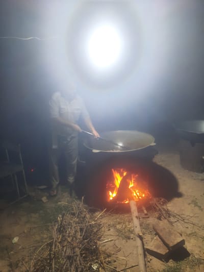 In the city of Vosobad, Tajikistan, a person is seen standing next to a large, dark pot, possibly cooking. The scene is illuminated by a bright, overhead light source that creates a halo effect and obscures details. A fire burns beneath the pot, casting a warm glow on the surrounding dry twigs and branches. The ground appears to be dirt or sand. A chair and another pot are visible in the background. The overall atmosphere is dimly lit and hazy due to the intense light and smoke.