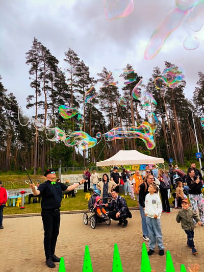 In Ulbroka, Latvia, an outdoor public event is captured under an overcast sky, featuring a bubble artist performing for a crowd. The main subject, a fair-skinned adult male, stands centrally on a light-colored paved area, wearing a black fedora, sunglasses, a black short-sleeved shirt with a bright green bow tie, black pants, and black shoes. He holds two long black rods connected by a string, actively generating numerous large, iridescent soap bubbles of various shapes and sizes that float upwards and across the scene, reflecting a spectrum of colors.
Behind the artist, a diverse crowd of adults and children observes the performance. Many individuals are looking upwards at the bubbles. A young girl in a white hoodie and blue jeans stands in the mid-foreground, intently watching the bubbles. To her right, a young boy in a green jacket and orange cap is in motion, appearing to run. Further back, a man crouches next to a baby stroller, while other adults and children are gathered on the paved area and a grassy slope.
The background is dominated by a dense forest of tall, slender pine trees. In the midground, a white event tent with partial text "VIR TU VI" is visible, along with small yellow and blue children's furniture on a grassy area to the left. Metal barriers line parts of the grassy slope. A black lamppost and blue road signs depicting a pedestrian crossing are visible on the right. Three bright green, cone-shaped markers are positioned in the immediate foreground on the paved surface.
