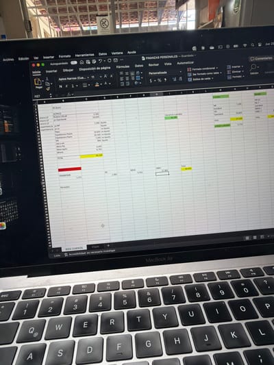 This image shows a close-up view of a MacBook Air laptop with an open spreadsheet document, likely a personal finance tracker. The spreadsheet displays columns and rows filled with financial data, including categories like "ACTIVOS", "PASIVOS", and amounts. The laptop is placed on a surface that is not fully visible but appears to be indoors. The background suggests an indoor environment with a view of a tiled structure and a barred window, potentially indicating a residence or office space. The screen is illuminated, and the keyboard is visible in the foreground. The image quality is slightly blurred due to the angle and focus. There are no people visible in the image. The scene does not provide any specific clues about the location in Cancún, Mexico, beyond the possibility of it being an indoor setting there.