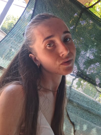 The media file is a close-up, sideways, over-the-shoulder shot of a young woman. She has long, dark brown hair, some of which is falling across her face and shoulder. She is wearing a white top and a small hoop earring in her visible ear. Her skin has a scattering of freckles across her nose and cheeks. The background consists of a dark green mesh netting, through which sunlight filters, illuminating dappled green foliage and what appears to be a bright sky beyond. The lighting suggests it is daytime, likely with bright sunshine. The woman's expression is neutral, with her gaze directed towards the camera. The scene is outdoors, possibly in a garden or patio area, given the netting and greenery. There is no visible text in the media file. The provided location context is Vypasne, Ukraine.