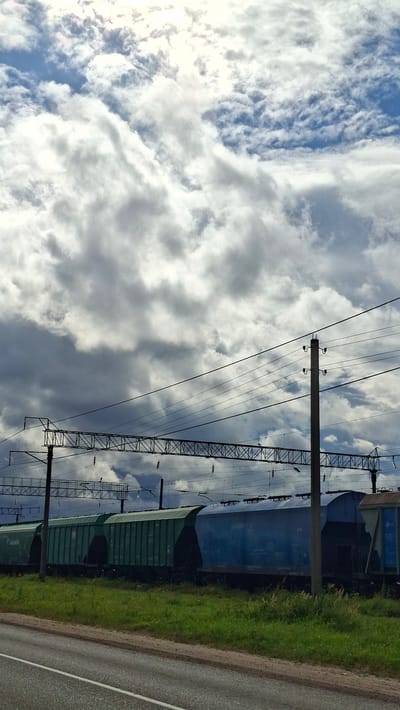 The image depicts a train on railroad tracks, likely located in the city of Smalyavichy, Belarus, as indicated. The train consists of multiple cargo cars, with some painted green and others blue. The train is positioned in front of a grassy area, with a paved road visible in the foreground. Overhead, a complex network of power lines and supports stretches across the frame, suggesting an electrified railway. The sky is filled with dramatic clouds, with patches of blue sky visible and the sun breaking through in the upper portion of the image. The overall impression is a somewhat industrial landscape under a dynamic sky.
