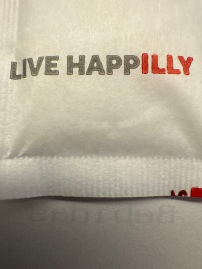 The scene, located in Andratx, Spain, depicts a close-up of a white, textured surface, likely a fabric or non-smooth paper material. Prominently displayed is the phrase "LIVE HAPPILY" in bold, sans-serif capital letters. The words "LIVE HAP" are dark grey, while the letters "PILLY" are rendered in red. Below this text, a distinct horizontal crease or seam is visible across the material. Further below the crease, faint, reversed grey text is partially discernible. Near the bottom right edge of the frame, several small, irregular red specks are present on the white surface. No people, actions, or interactions are visible. The environment appears to be an interior setting based on the uniform, diffuse lighting.