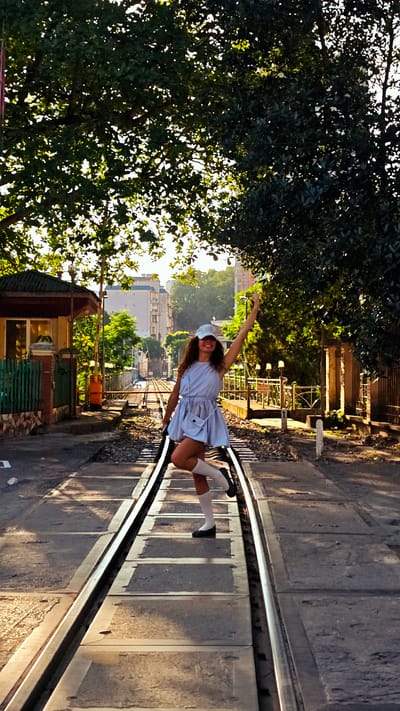 A young woman in a light blue dress and white cap is standing on a railroad track in Lao Cai Ward, Vietnam. She is posing with one arm raised and one leg bent. The railroad track runs down the center of the frame, leading towards a building in the distance. There is a small guardhouse to the left and a fence to the right of the tracks. Trees overhang the area, casting shadows. The lighting suggests it is either early morning or late afternoon.
