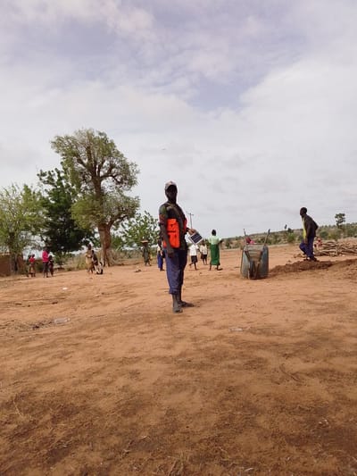 The image shows a scene in Mulke Barikin Sayejo, Nigeria. The ground is dry and reddish-brown. Several people are present, including a man in the foreground wearing blue pants, sandals, and an orange vest. He is holding a solar panel. In the background, there are other people, trees, a wheelbarrow, and some piles of wood. Some people are gathered near a building in the distance. The sky is cloudy.